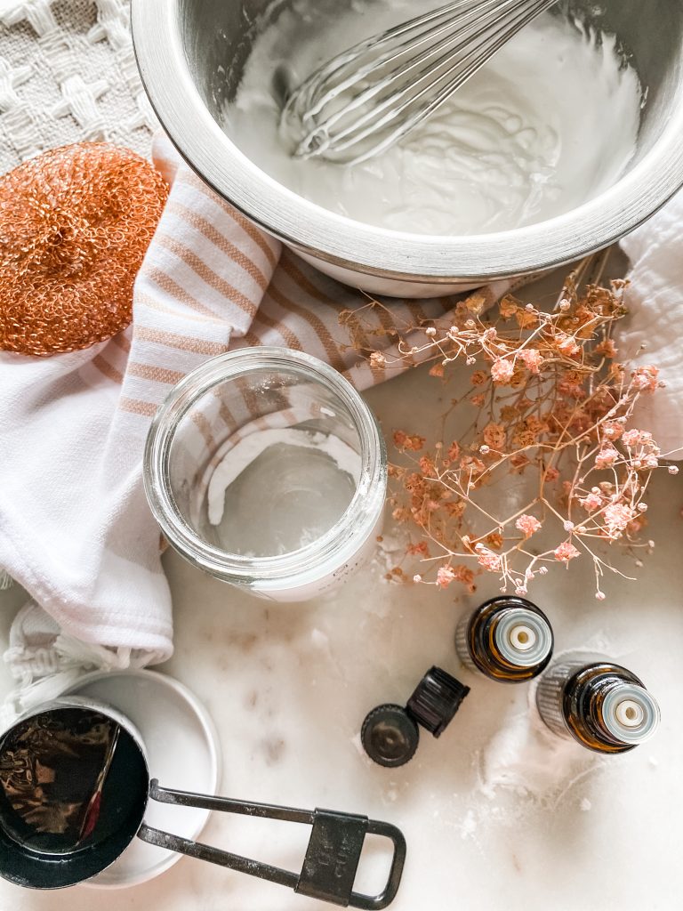 Mixing bowl with ingredients glass jar essential oil bottle measuring cup small towel and rose colored babies breathe