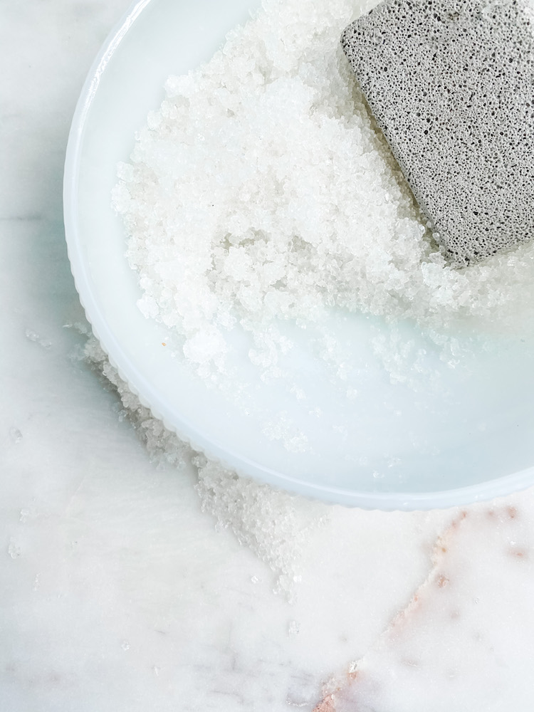 foot scrub in a white bowl with grey scrubber all on a marble table