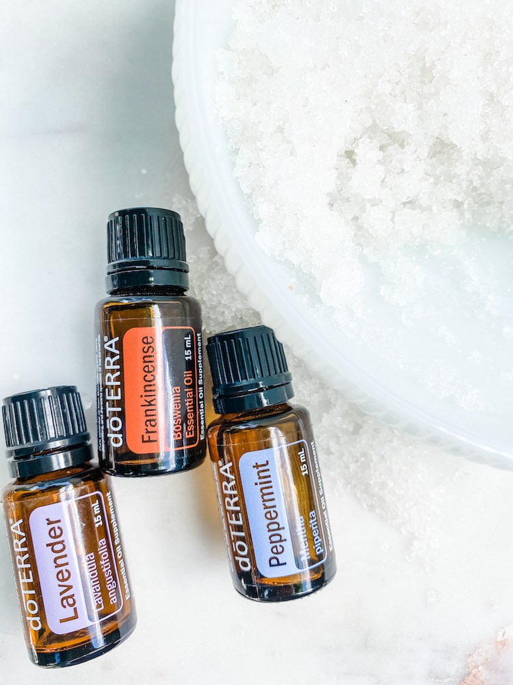 foot scrub in a large white bowl with 3 bottles of essential oils laid next to it on top of a marble background