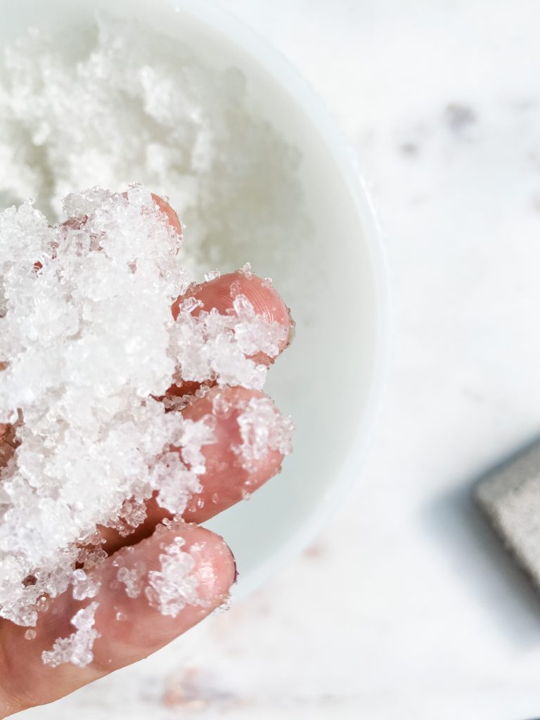close up of the tip of a hand holding white foot scrub with a bowl of foot scrub blurred in the background