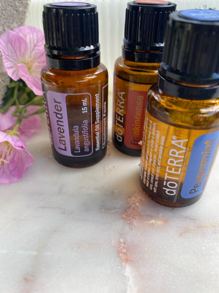 close up of essential oils- lavender, peppermint, and frankincense on marble with flowers next to them