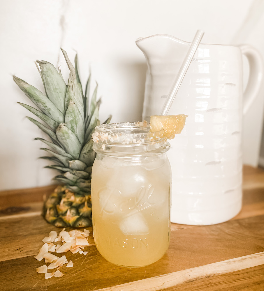 summer drink with pineapple with coconut crips pineapple top and pitcher shown on a cutter board