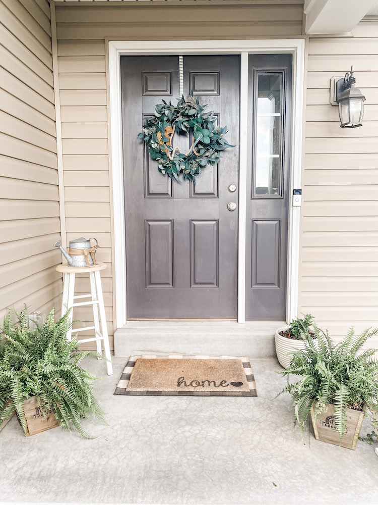 small front porch with 2 ferns, stool, watering can, and wreath on a dark grey door with a welcoming mat on the ground