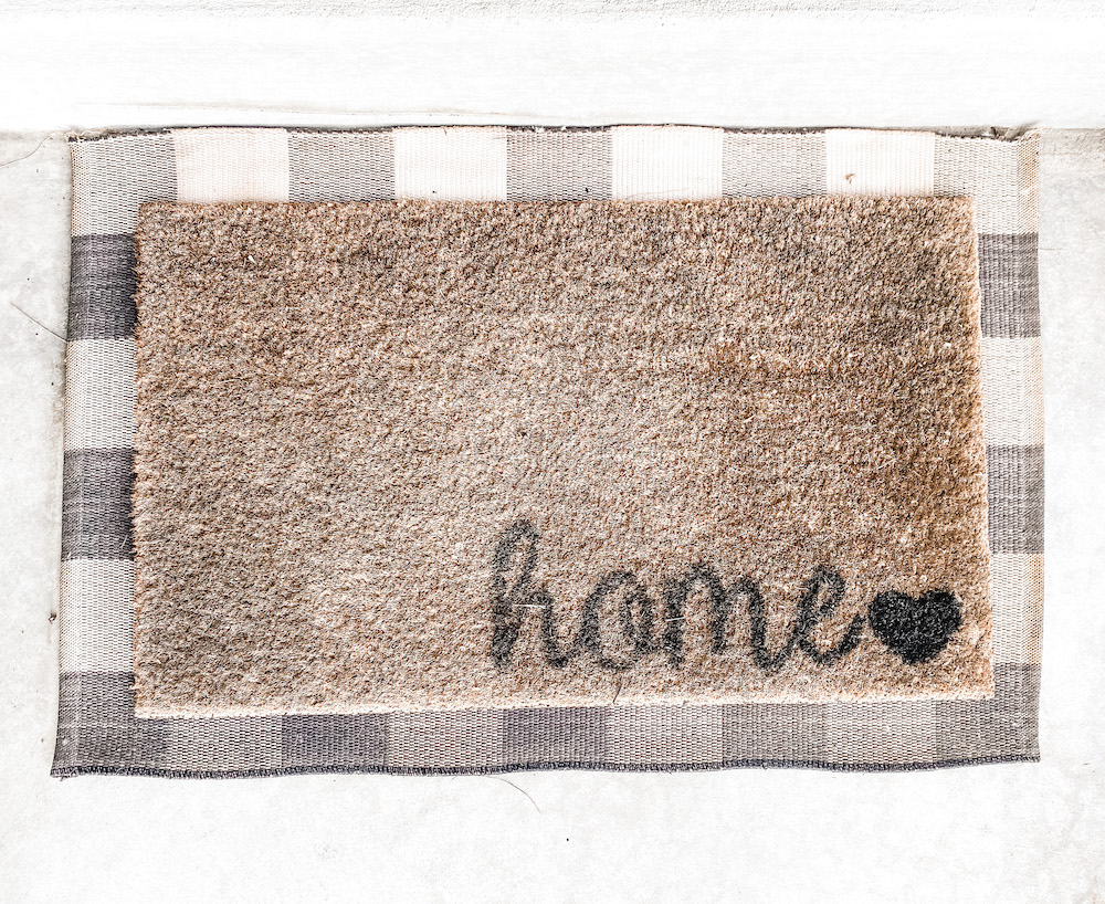 Brown welcome mat with the word home on it laid on top of a black and white buffalo plaid rug