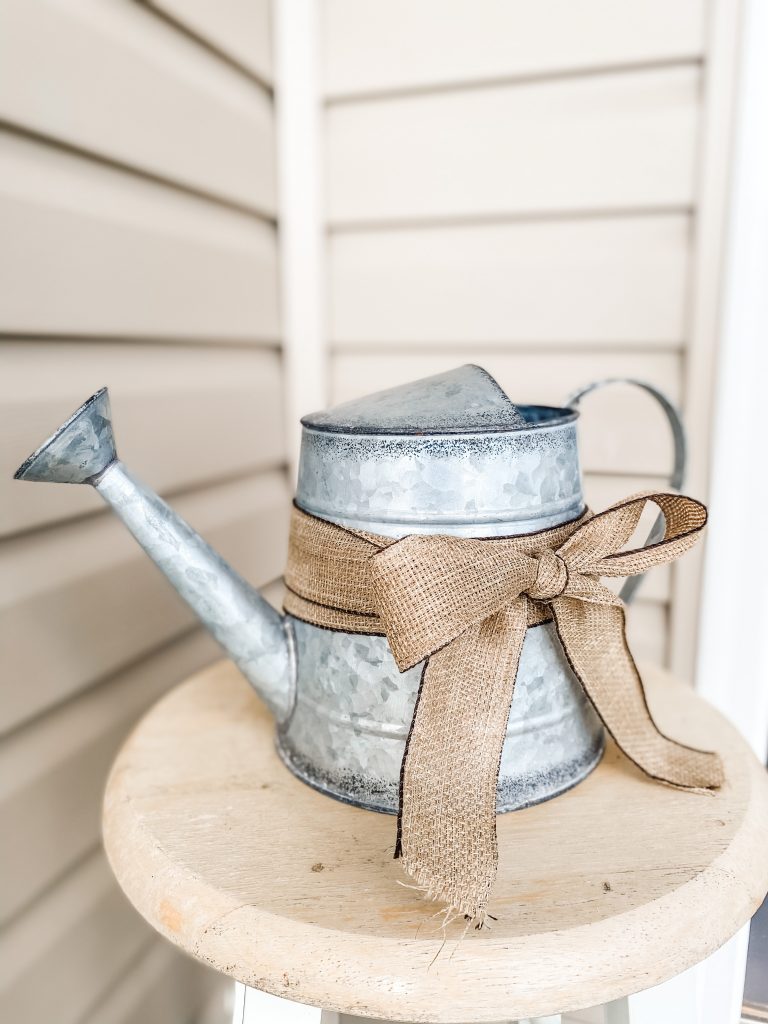 a galvanized watering can with a jute ribbon sitting on top of a raw rood stool with tan siding in the background
