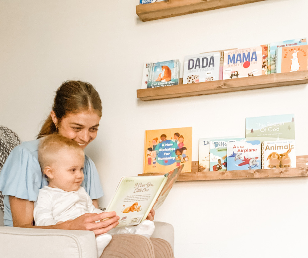 mom reading book to baby in a chair with books in the background on wall shelves