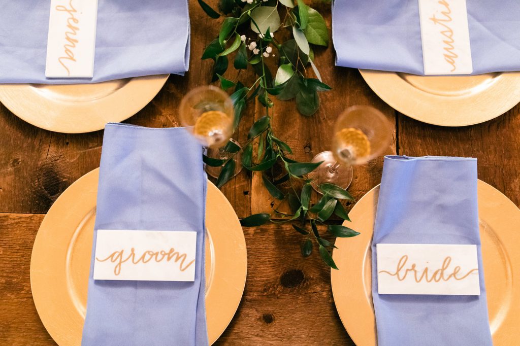4 place settings with gold chargers, dusty blue napkins, and marble tile on top with name all on wooden surface with greenery runner