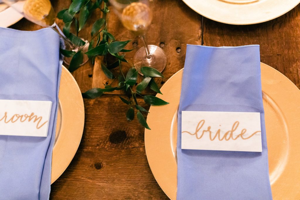 table setting with gold chargers and periwinkle napkins with bride and groom marble name plates on wood table