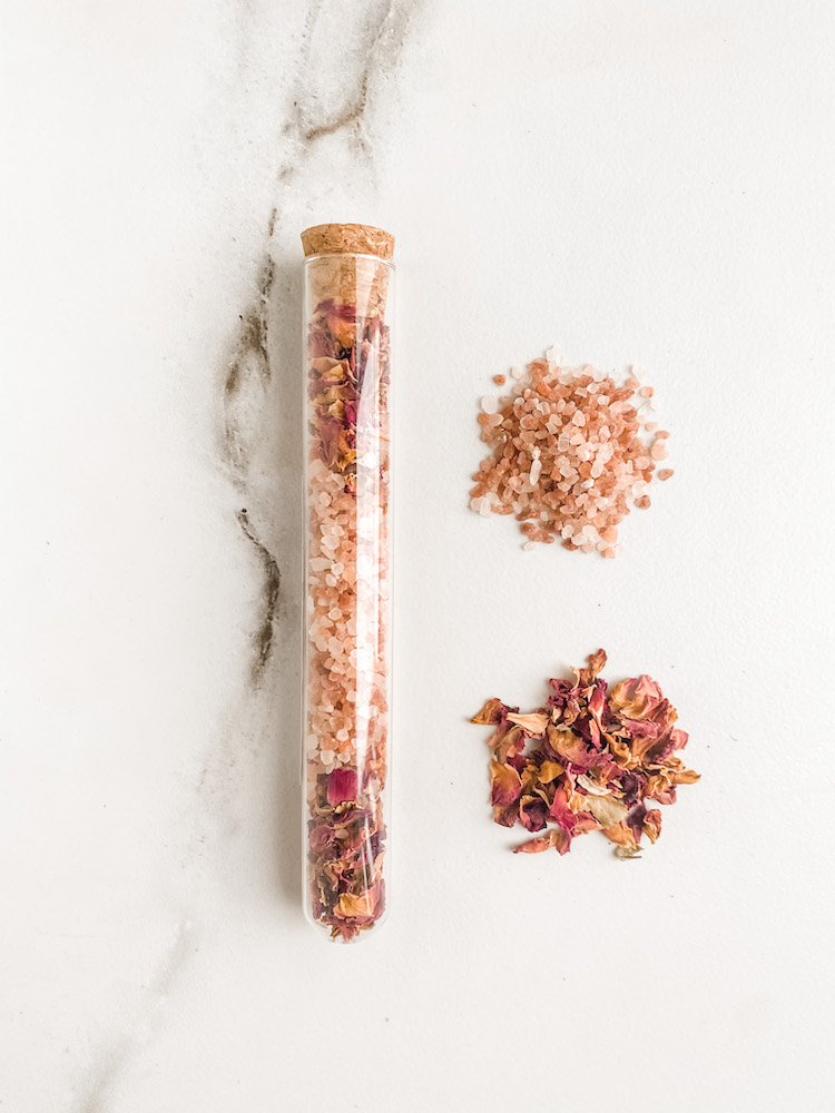 glass tube filled with dried roses and pink salt with the same ingredients separated in piles beside it set on a white surface