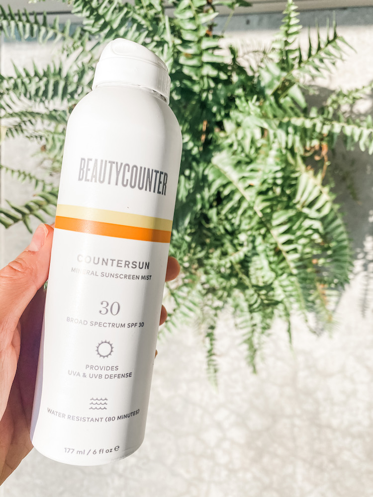 hand holding Beautycounter spray countersunk sunscreen above of a fern