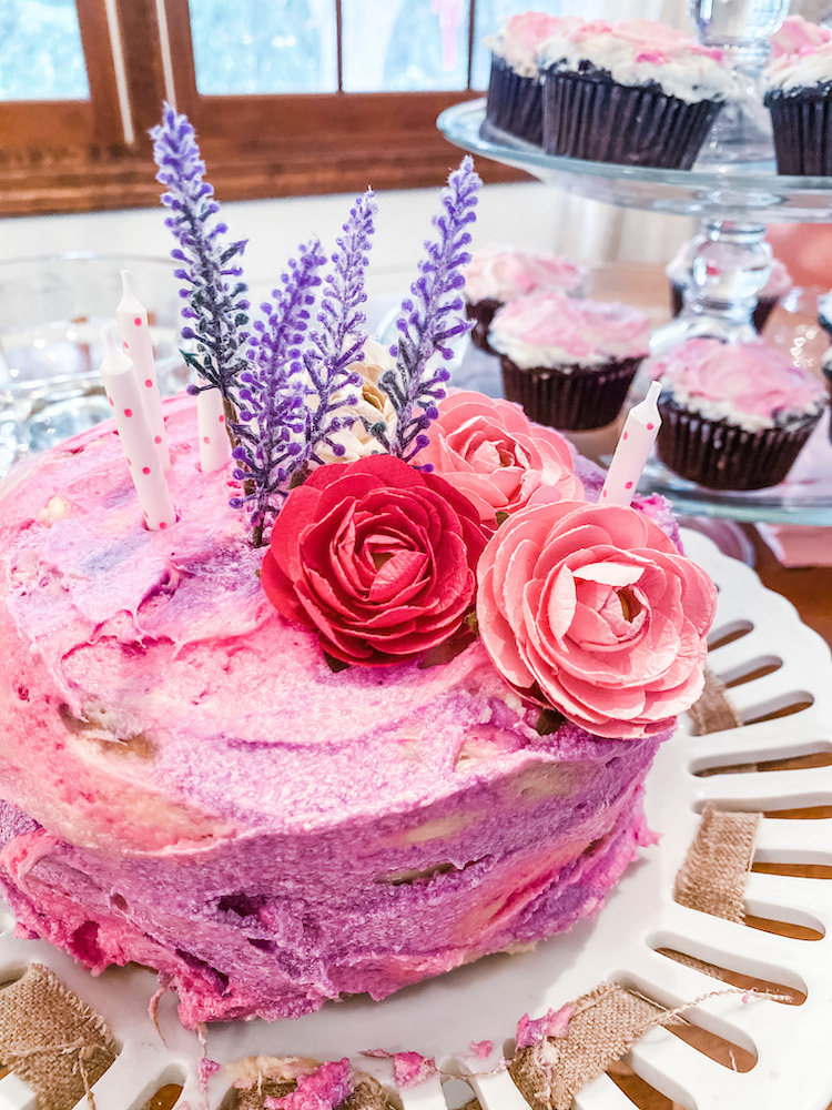 a cake with purple and pink flowers and purple and pink marbled icing on a white cake stand with chocolate cupcakes in the background