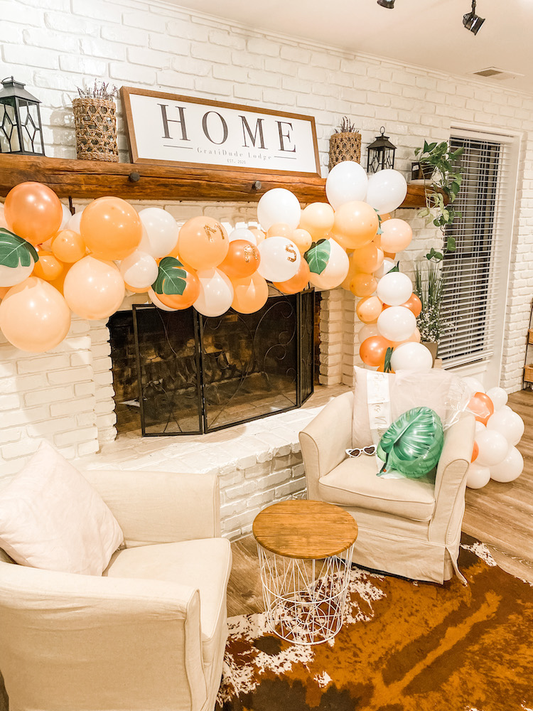 peach and white balloon garland with faux banana leaves hanging on a white fireplace with a home sign above it, and cozy chairs in front