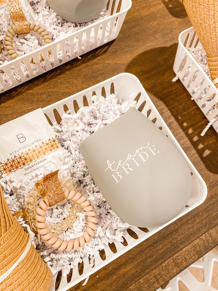 close up of a gift basket with a rolled up straw hat, Beautycounter sunscreen, hair ties, and silicone gray team bride cup with white shredded paper inside the basket